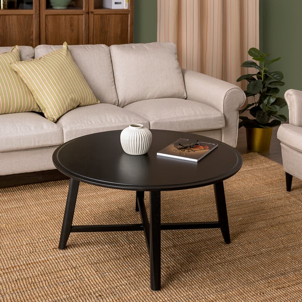 Small round black coffee table with a white vase and book, placed in front of a light-colored sofa with a green striped pillow, on a woven rug.