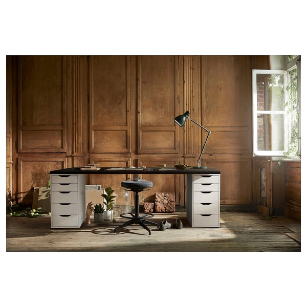 A sleek, white office setup featuring the LAGKAPTEN desk with drawers, a modern black chair, lamp, and potted plants, set against wooden panel walls.