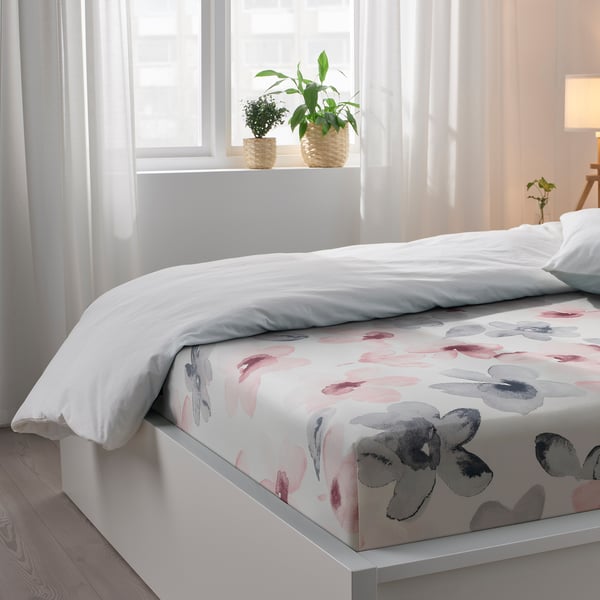 Bright bedroom with white bed, grey & pink floral sheets, large window with sheer curtains, serene & inviting.
