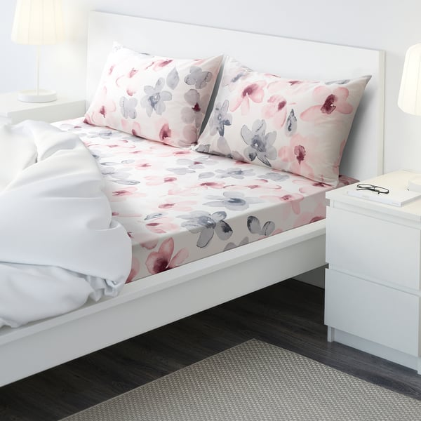 Bright bedroom with white bed, pink & grey floral sheets, white comforter, bedside table & lamp.