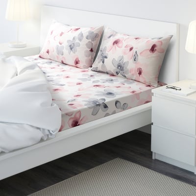Bright bedroom with white bed, pink & grey floral sheets, white comforter, bedside table & lamp.
