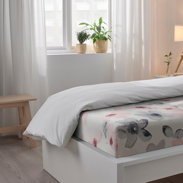 Cosy bedroom with white bedding, colourful floral sheet, wooden bench, and potted plants near a window with sheer curtains.