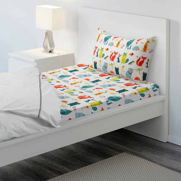 Colourful childs bedsheet with matching pillow on white bed, lamp on bedside table.
