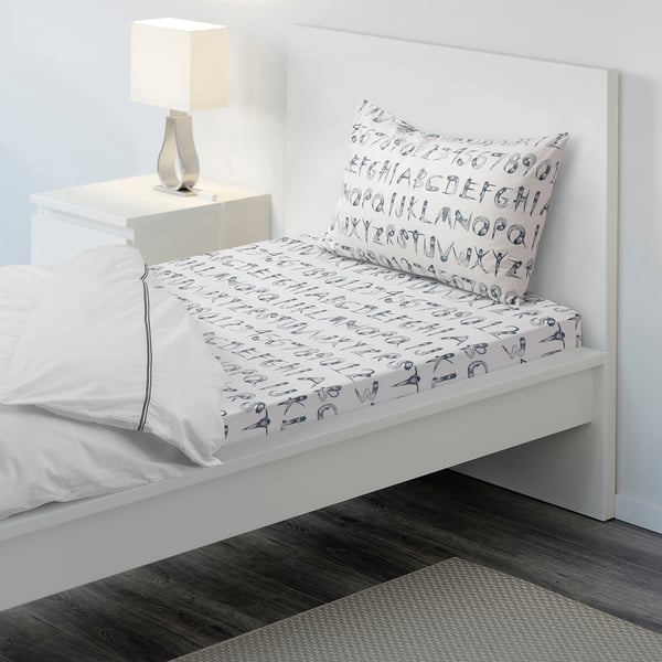 Small bed with white bedding, blue playful print, wooden floor, lamp.