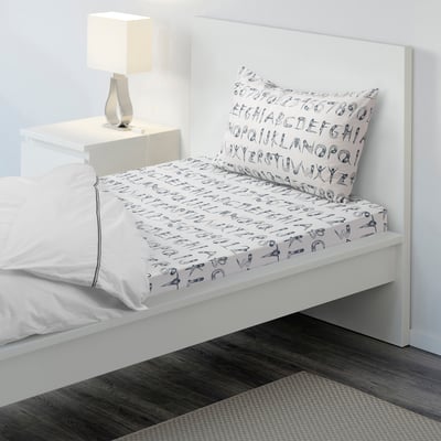 Small bed with white bedding, blue playful print, wooden floor, lamp.