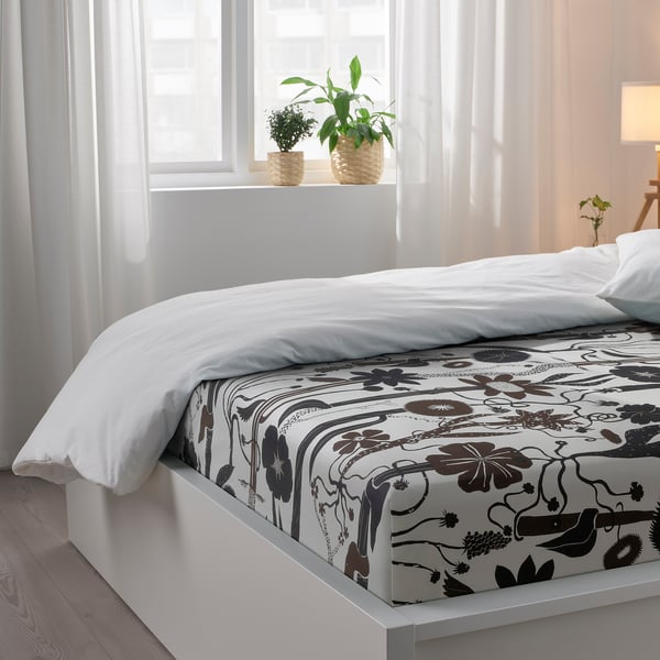 Cosy bedroom with floral bedspread, pillows, plants, and sunlight through curtains.