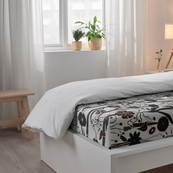 Bedroom scene: white bed with floral sheets, wood bench, potted plants near window with sheer curtains.