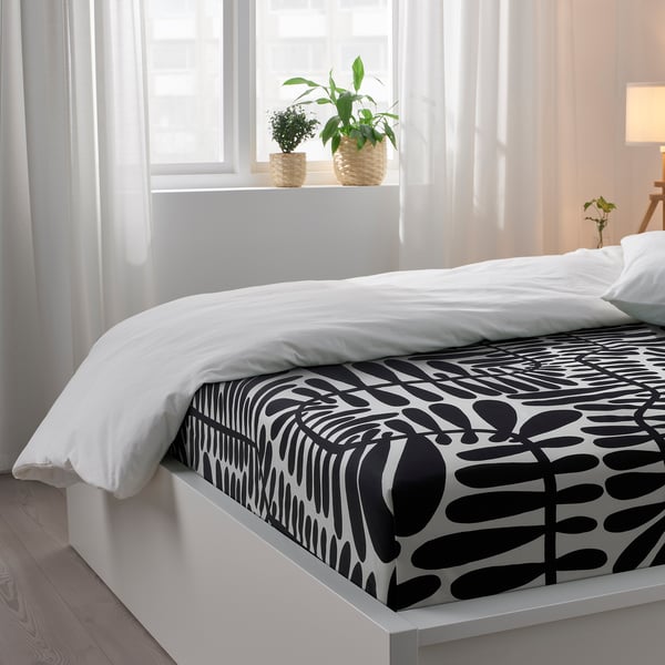 Bedroom scene with white bedding, black leaf-patterned sheet, and potted plants on windowsill.