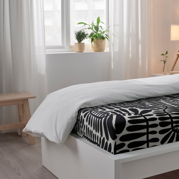 Bedroom with white bed, black and white bedsheet, two potted plants on windowsill, and a bench.