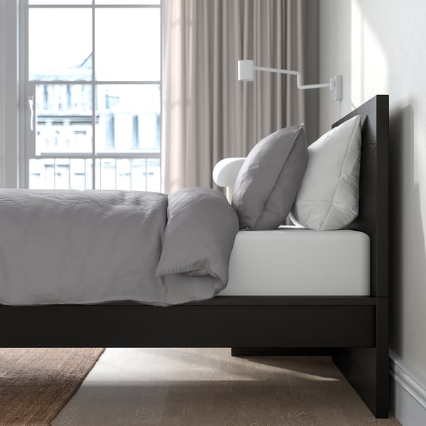 Bedroom scene with a modern black bed frame, grey bedding, and large pillows against a white headboard. A wall-mounted lamp illuminates the side.