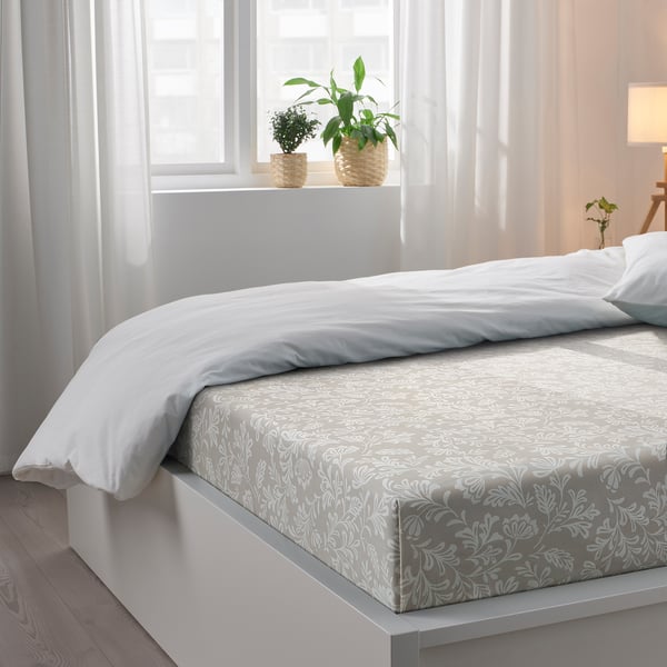 Bed with white linens, leafy print sheet, pillows, plants on windowsill, sheer curtains, wooden floor.