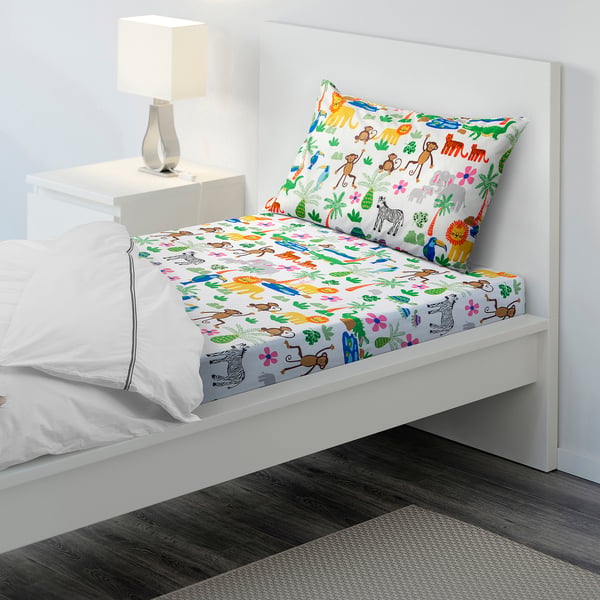 A small, single bed with NATTHÄGER colourful jungle animal sheets and a white pillow. The lamp beside it highlights the cosy setup.