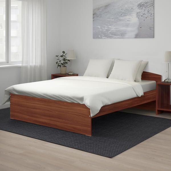 Minimalist bedroom with white bedding, large brown NODELAND bed, two small bedside tables, lamps, and a plant.