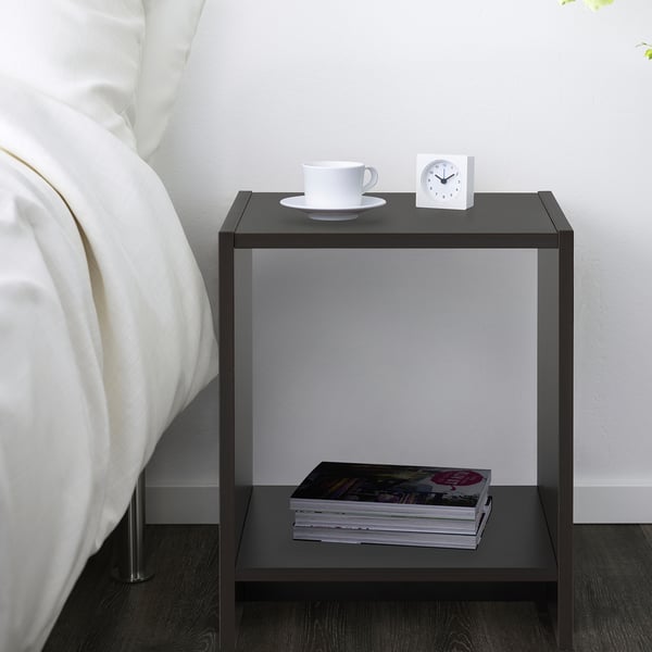 A black bedside table with a stack of magazines on the open shelf and a white alarm clock and cup on top, positioned beside a bed with white pillows.