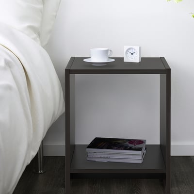 A black bedside table with a stack of magazines on the open shelf and a white alarm clock and cup on top, positioned beside a bed with white pillows.
