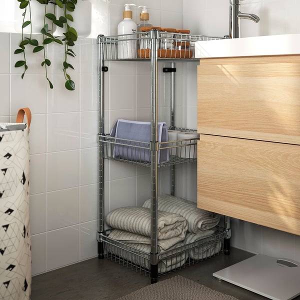 Modern bathroom with silver OMAR storage shelf. Holds folded towels and bottles, positioned beside a light wooden vanity and white tiled wall.