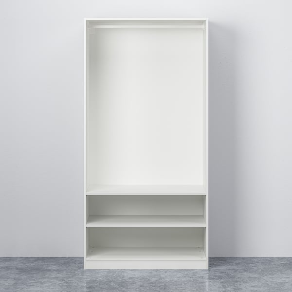 An empty, white, two-shelf PAX storage unit standing against a plain wall. It has adjustable feet and requires wall mounting.