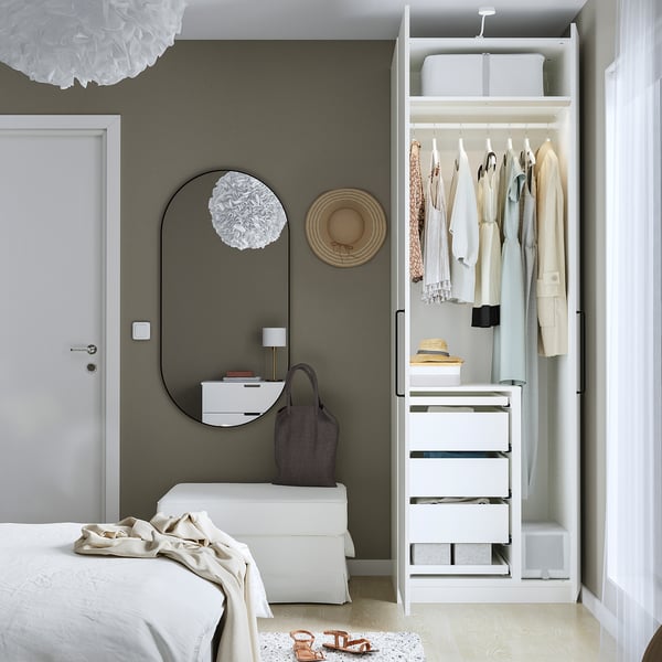 Bedroom with open wardrobe displaying clothes, drawers, and storage boxes. Mirror, lamp, and hat on wall; bed with white blanket nearby.
