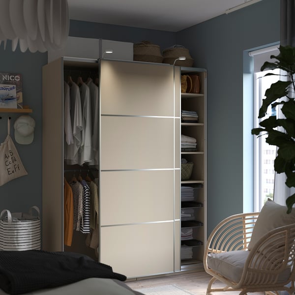 Modern bedroom with beige PAX wardrobe, sliding doors, hanging clothes, shelves, baskets, and a wicker chair.