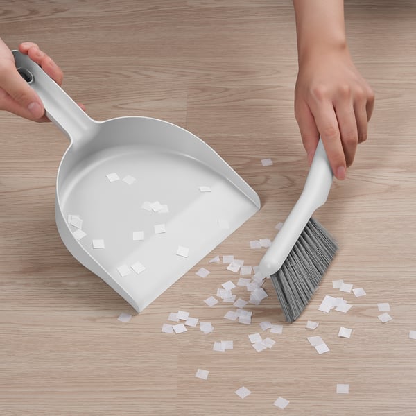 Person uses PEPPRIG dustpan and brush to sweep up small squares on wooden floor. Brush is snapped onto dustpan handle.