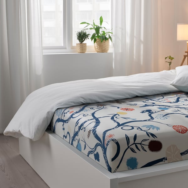 A bed with floral bedsheet, white, blue-red flowers, white pillow. Bedside window with white curtains and two plants.