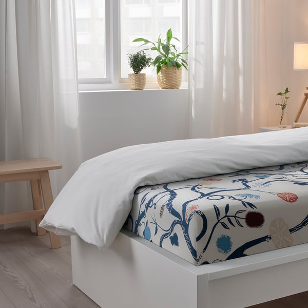 Bedroom with white bedspread featuring floral print, two potted plants on window sill, wooden stool, and sheer curtains allowing natural light.