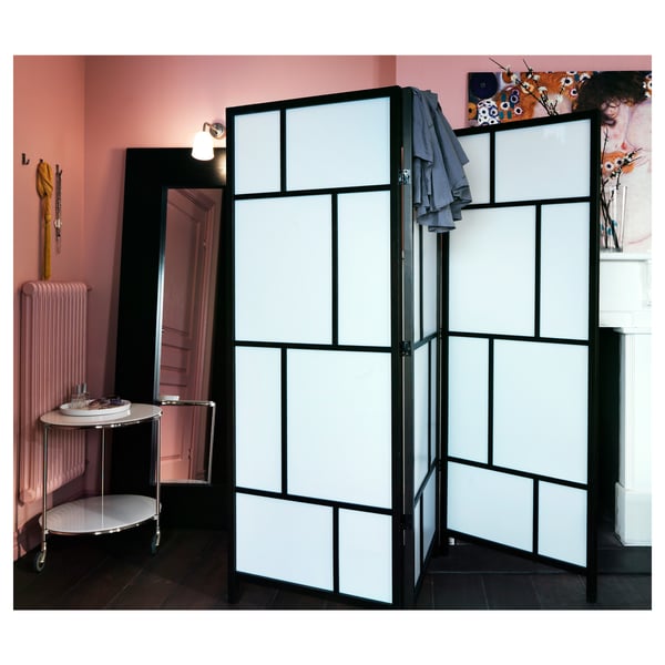 Black room divider with frosted panels creates distinct spaces, positioned in a pink room with a side table, a radiator, and wall decorations.