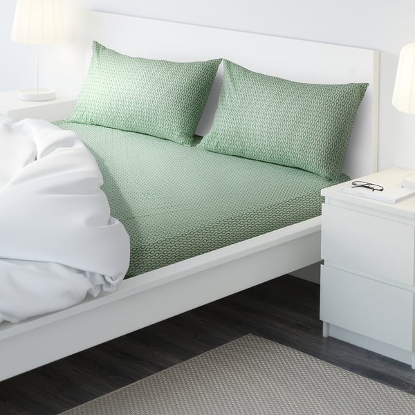 A modern bed with green patterned sheets, two white pillows, a white blanket, and a bedside table with a lamp.