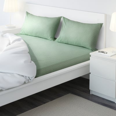 A modern bed with green patterned sheets, two white pillows, a white blanket, and a bedside table with a lamp.