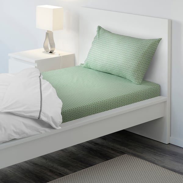 A neatly made bed with green patterned sheets and a white pillow. The lamp beside suggests a cosy and inviting sleeping area.