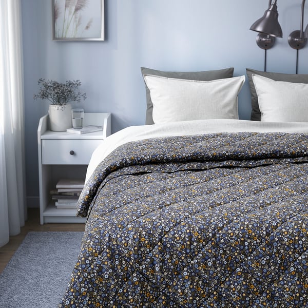A cosy bedroom with a floral-patterned comforter on a bed, white pillows, and a side table with books and a vase.