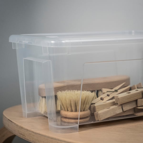 Clear plastic box with wooden clothes pegs and brushes inside, sitting on a light wooden shelf.
