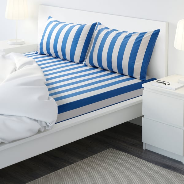 Striped bluesheet, white walls, side lamps, bedside table with book, dark rug.