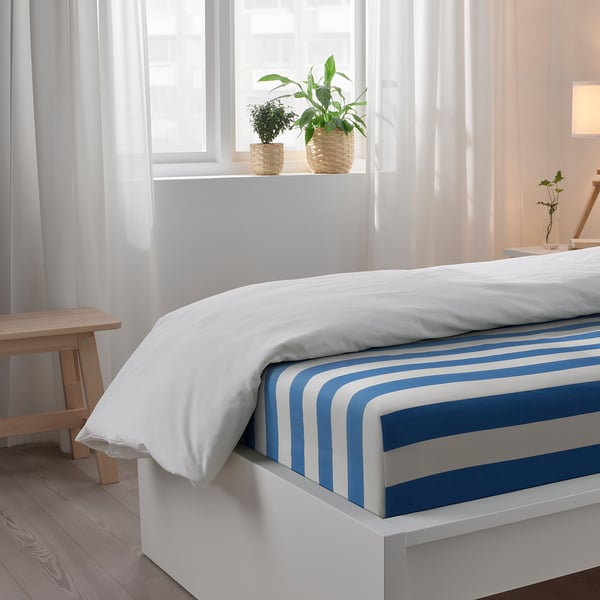 Bedroom scene with white bed, blue-striped sheets, white pillows, wooden stool, and potted plants on windowsill.