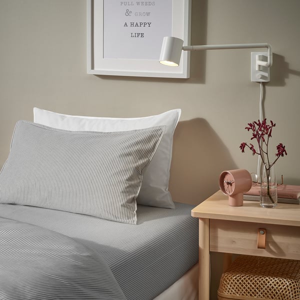 Bedroom with SOLFIBBLA striped grey pillows, wooden bedside table, white wall light, and vase with red flowers.