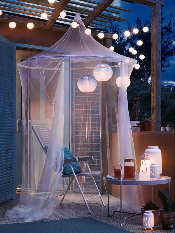 An outdoor relaxation space with SOLIG net canopy, white hanging lights, two chairs, a table with beverages, plants, and wooden pergola.