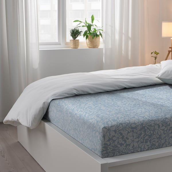 A crisp white bed with a blue floral-patterned sheet set, featuring soft cotton/lyocell fabric, lies beneath a window with potted plants.