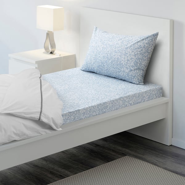 Twin size bed with white and blue floral sheets, crisp cotton, with a neat lamp on a small bedside table beside it.