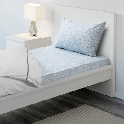 Twin size bed with white and blue floral sheets, crisp cotton, with a neat lamp on a small bedside table beside it.