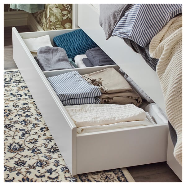 White SONGESAND bed frame with drawers, folded linens in blue and beige, classic rectangle with rounded edges and high legs.
