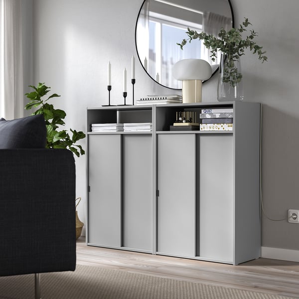Grey storage unit with open/closed sections, lamp, mirror, plant, and surrounding large plant & sofa.