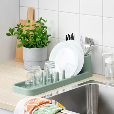 Kitchen sink with green STÄMLING dish drainer holding plates, glasses, and cutlery; plant and chopping board nearby.