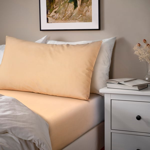Neatly made bed with beige bedding, wooden frame, laptop & vase on white bedside table, hillside picture above.