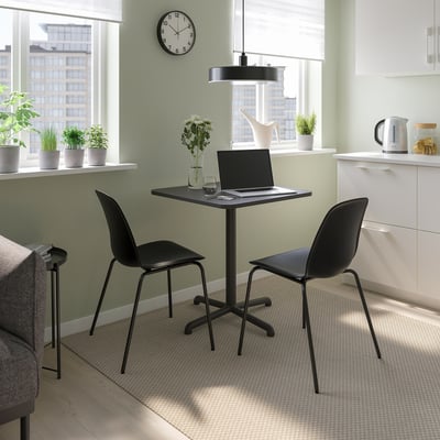 Small home office with STENSELE black table & chairs, laptop, plants, white wall, large window, kitchenette.