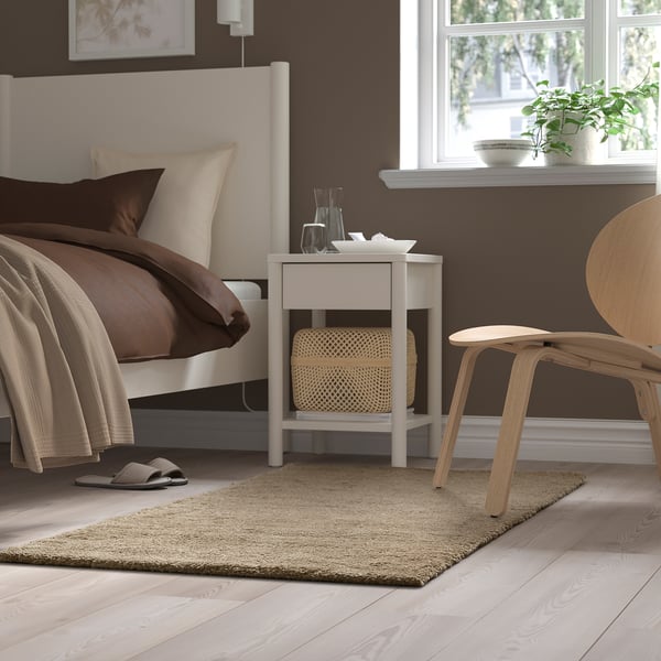 Bedroom with a brown bed, white bedside table, small white rug, light wood chair, and a window with plants.
