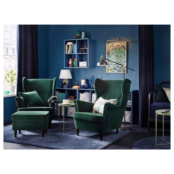 Living room with two green velvet armchairs, footstool, blue walls, dark curtains, rug, and shelves with decorative items.
