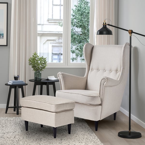 Modern living room: beige wing chair & ottoman, black side tables, lamp. Cosy, neutral tones.