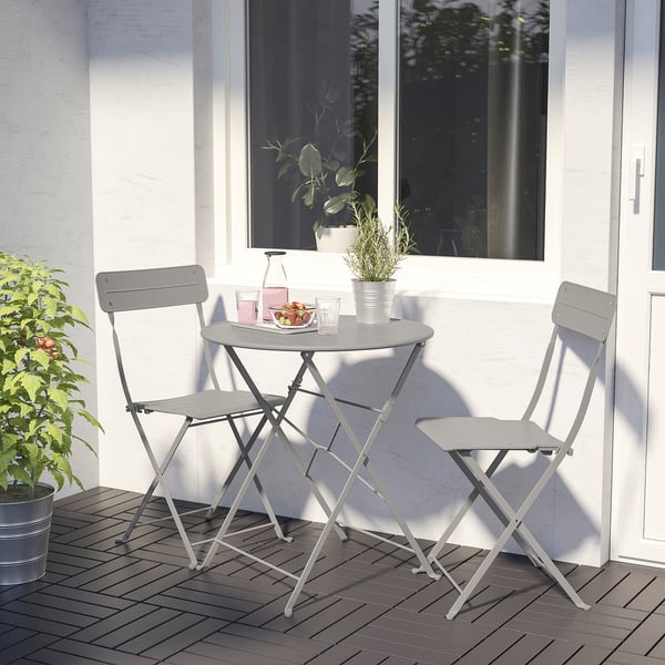 Small grey foldable patio set with round table and chairs. Perfect for compact spaces, placed near a white wall. Chairs fold flat for easy storage.