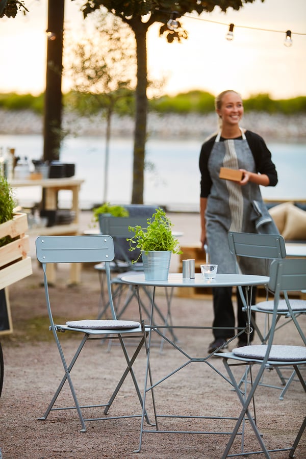Outdoor dining area with blue metal chairs and round table. Person in apron, potted plant on table, serene lake background.