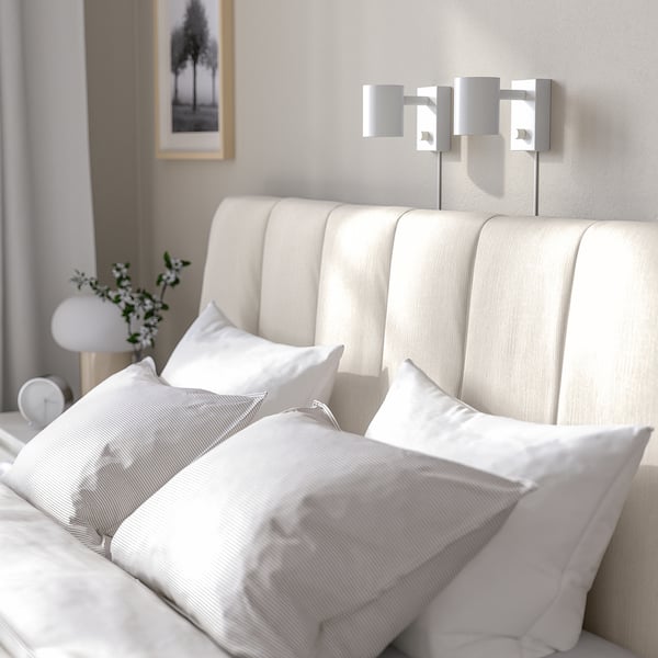 Bed with beige striped headboard, two wall-mounted lights, white pillows, and vase with greenery.
